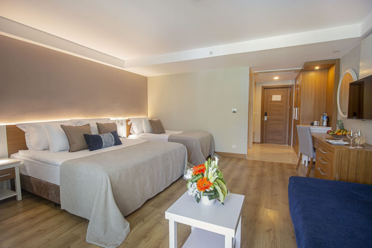 A hotel room with two beds, a desk, a chair, a small table with a vase of flowers, and a mirror on the wall.
