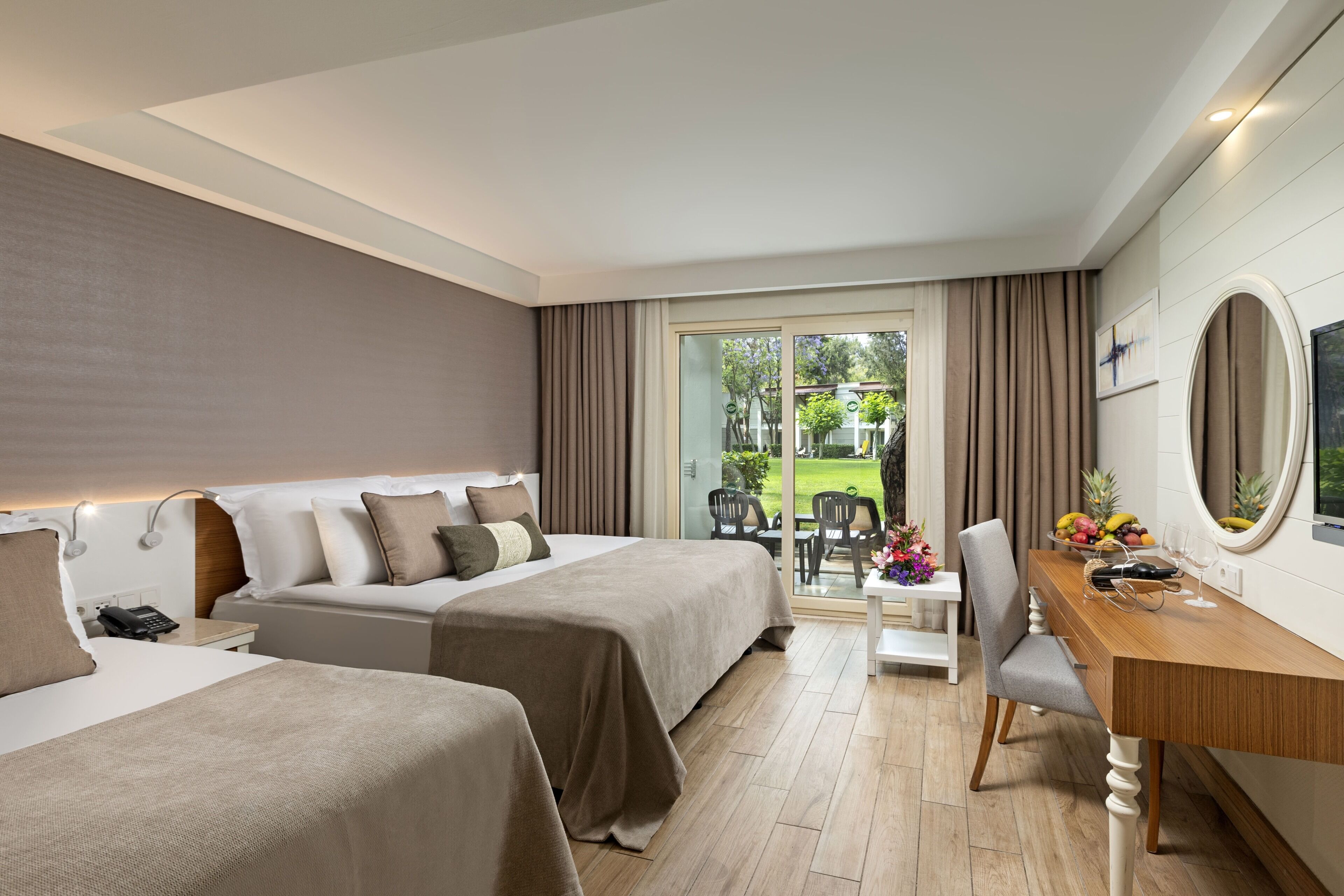 A hotel room with two beds, a wooden desk, a chair, a dining table, a telephone, and a large window with curtains.