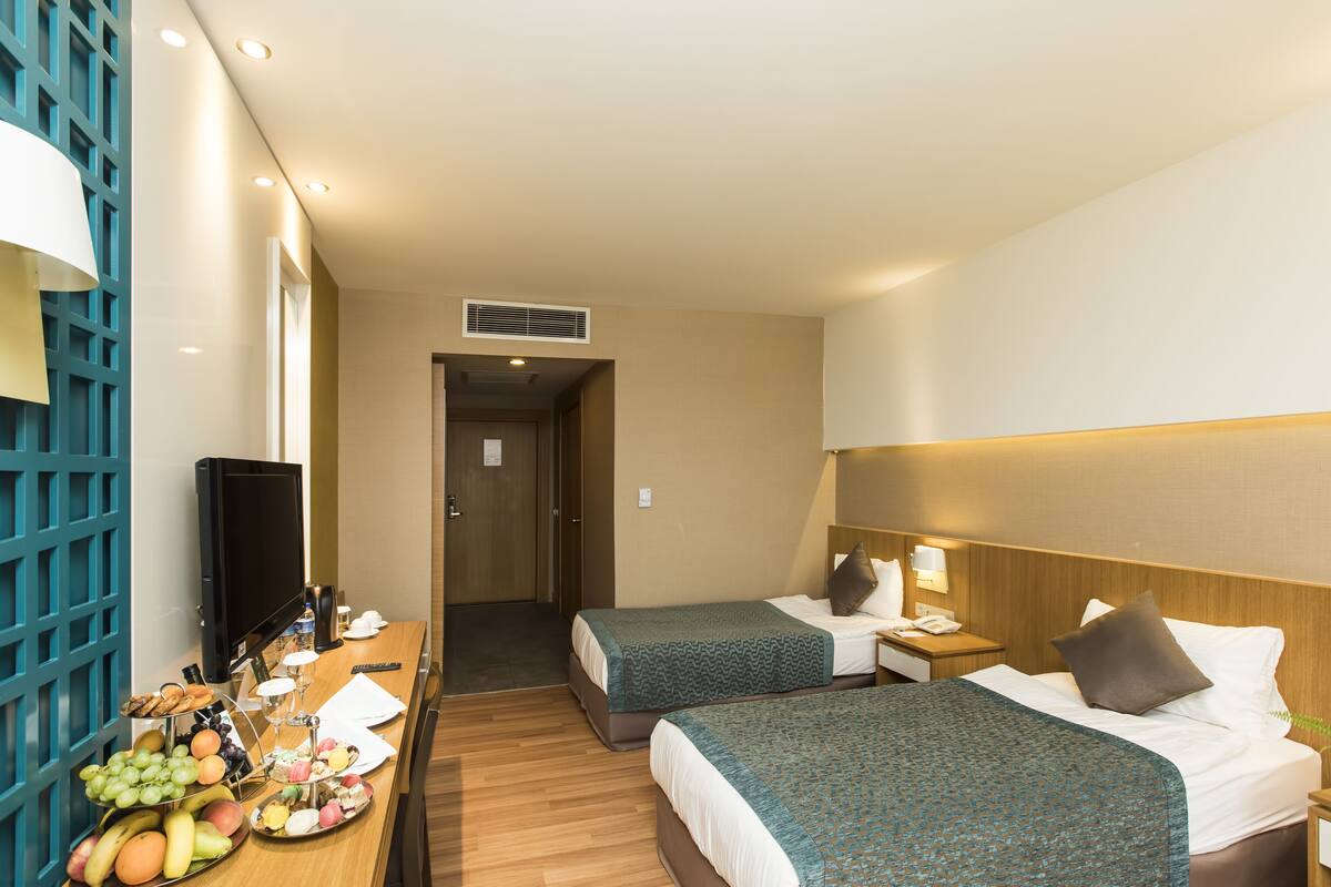 A hotel room with two beds, a TV, a desk with a fruit basket, and a bathroom visible in the background.