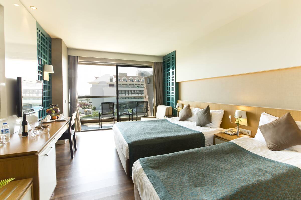 A hotel room with two beds, a desk with a chair, a small table, and a balcony with a view of buildings.