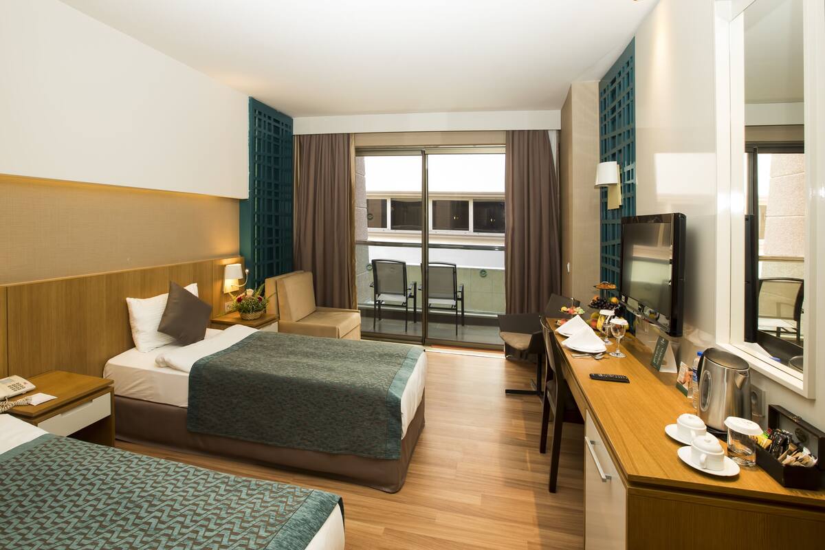 A hotel room with two beds, a desk with a coffee maker, a TV, and a balcony with a view of the city.