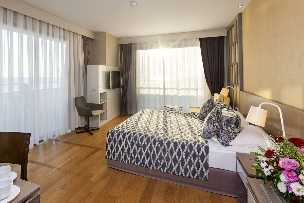 A hotel room with a bed, a desk with a kettle, a chair, a TV, and a window with curtains.