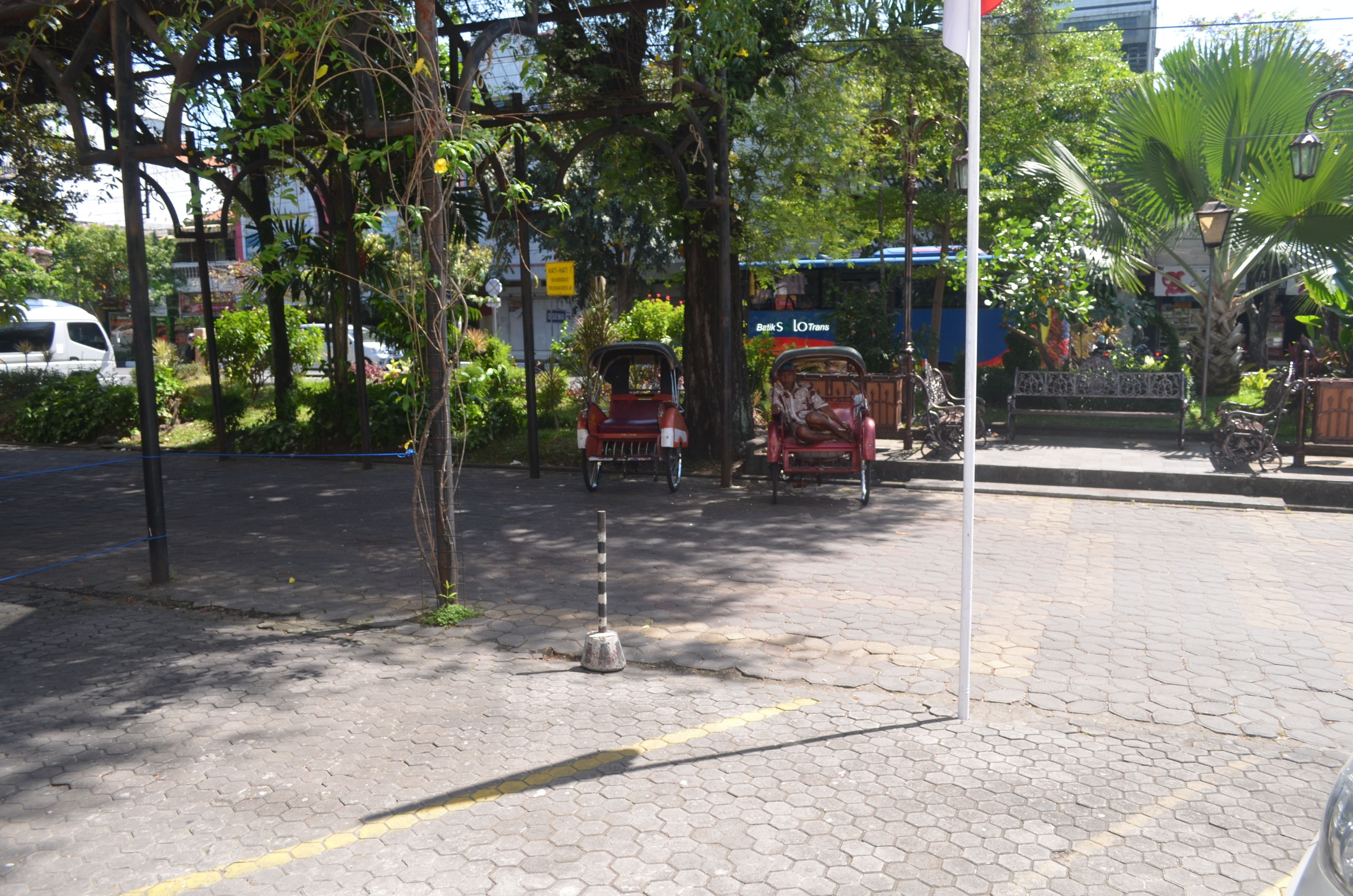 Una calle pavimentada con una banca, un tuk-tuk y un coche estacionado.