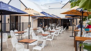 Un café en plein air avec des chaises en métal blanc et des tables en bois, à l’ombre de parasols arborant la marque « BUMBERS ».