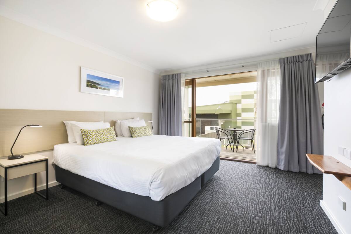 A hotel room with a large bed, a desk with a lamp, a view of the outside balcony, and a painting on the wall.