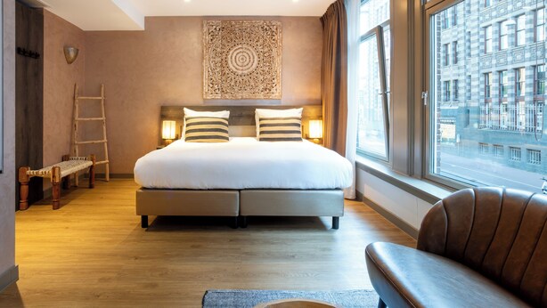 Habitación de hotel moderna con una cama grande, un banco, un sofá y vistas de la ciudad a través de amplias ventanas.