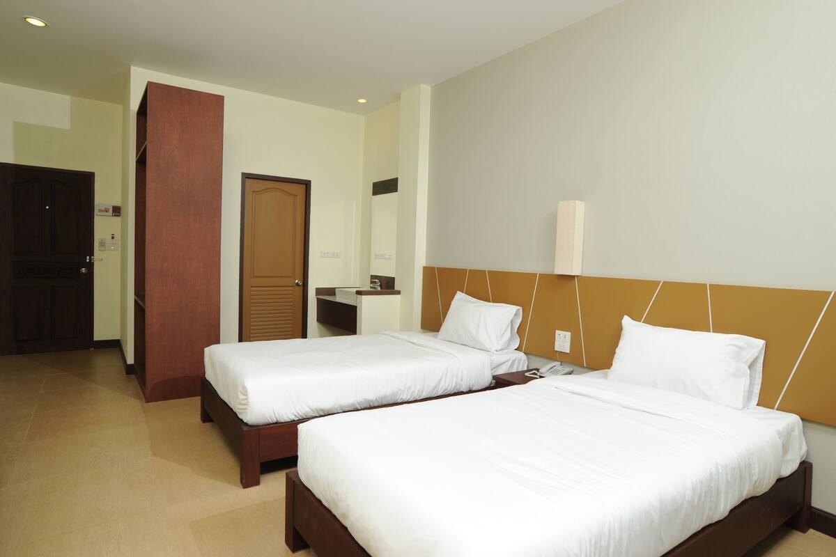 A hotel room with two single beds, a wooden headboard, a bedside table with a lamp, and a door leading to another room.
