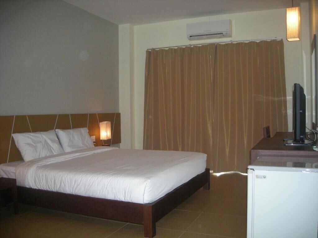 A hotel room with a bed, a nightstand with a lamp, an air conditioning unit, a television, and a refrigerator.