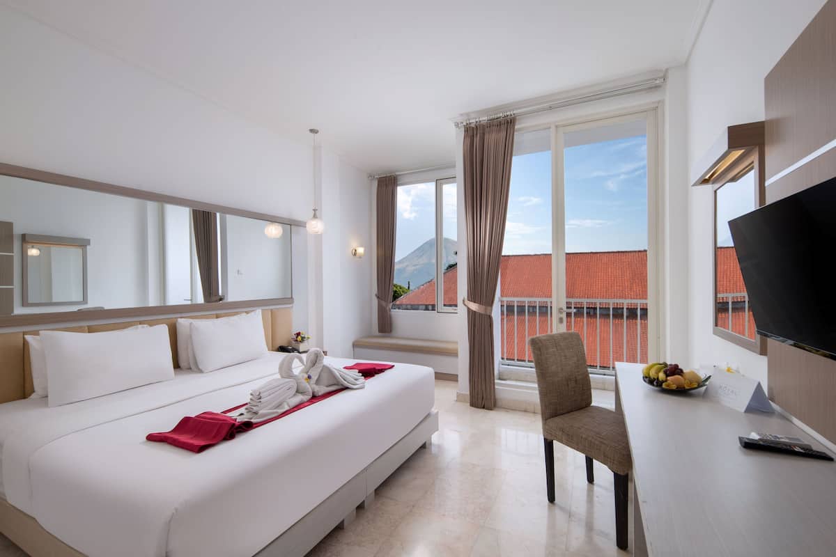 A hotel room with a bed, a desk with a fruit bowl, a chair, a TV, and a view of a mountain.