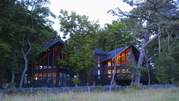 A large, dark wooden cabin with multiple windows and balconies, surrounded by trees. - Brockwood Hall (Millom)