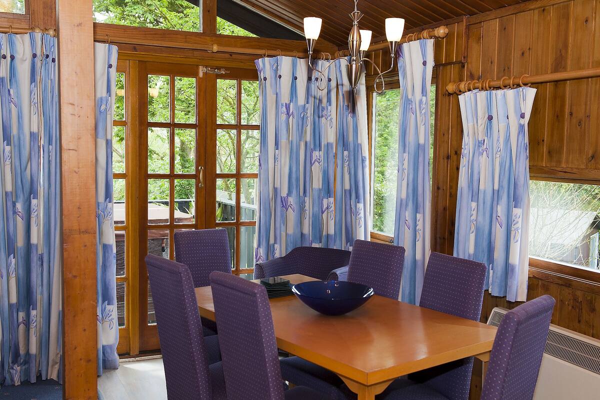 Un comedor de madera con mesa y sillas, ventanas amplias con cortinas y vistas al exterior.