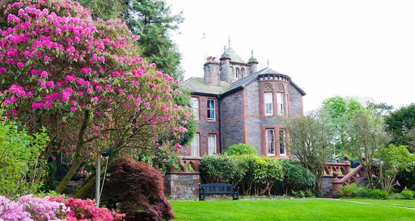 A large stone building with a turret and a well-maintained garden with pink flowers.