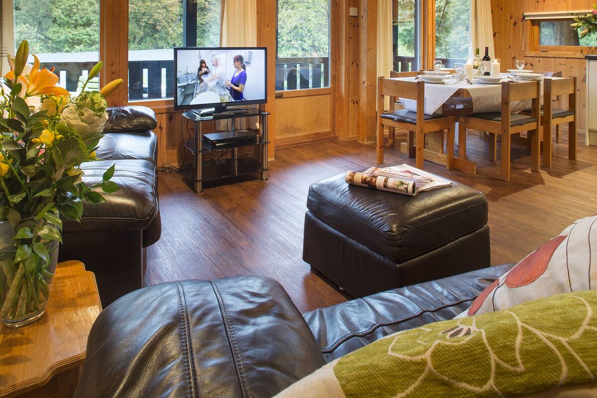 Una sala de estar acogedora con un sofá de cuero, una mesa de comedor preparada para cuatro personas y un televisor mostrando una escena.