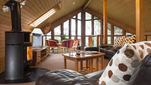 A cozy cabin interior with a wood-burning stove, leather seating, a dining area, and large windows offering natural light and views. - Brockwood Hall (Millom)