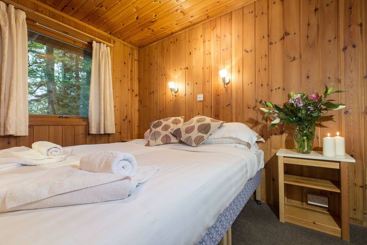 Una habitación de cabaña de madera con cama, toallas, una mesita de noche con velas y una ventana con cortinas.