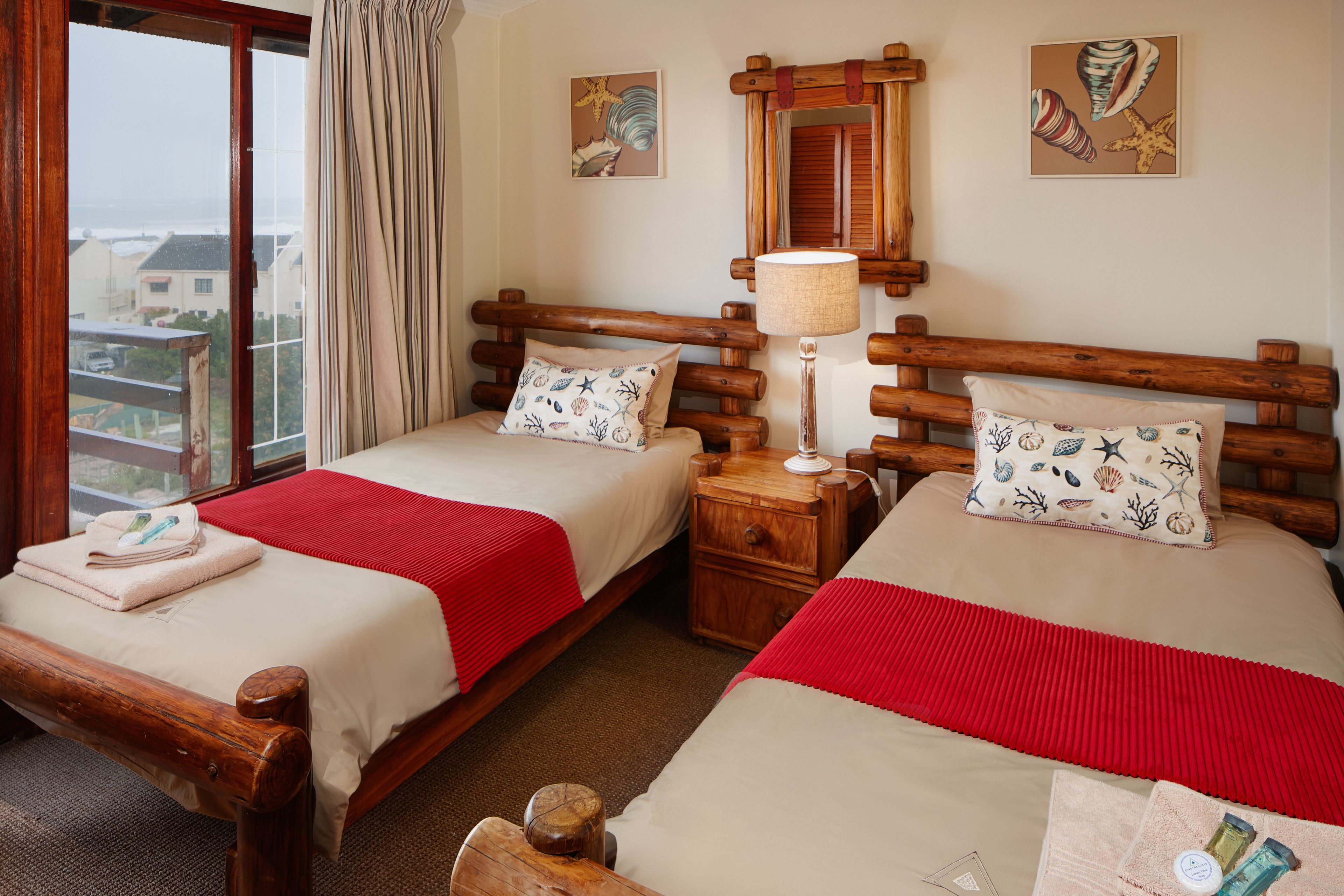 A hotel room with two wooden beds, a red blanket, a wooden nightstand, a lamp, and a window with a view of buildings.