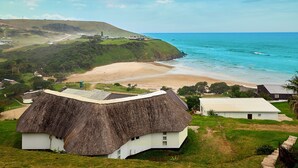 A thatched-roof house with a white exterior, situated on a grassy hill overlooking a beach and ocean. - First Group Hole in the Wall (King Sabata Dalindyebo)