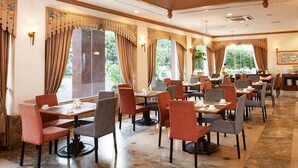 Een ruime eethoek met meerdere tafels en stoelen, grote ramen met gordijnen, en een centrale kroonluchter.