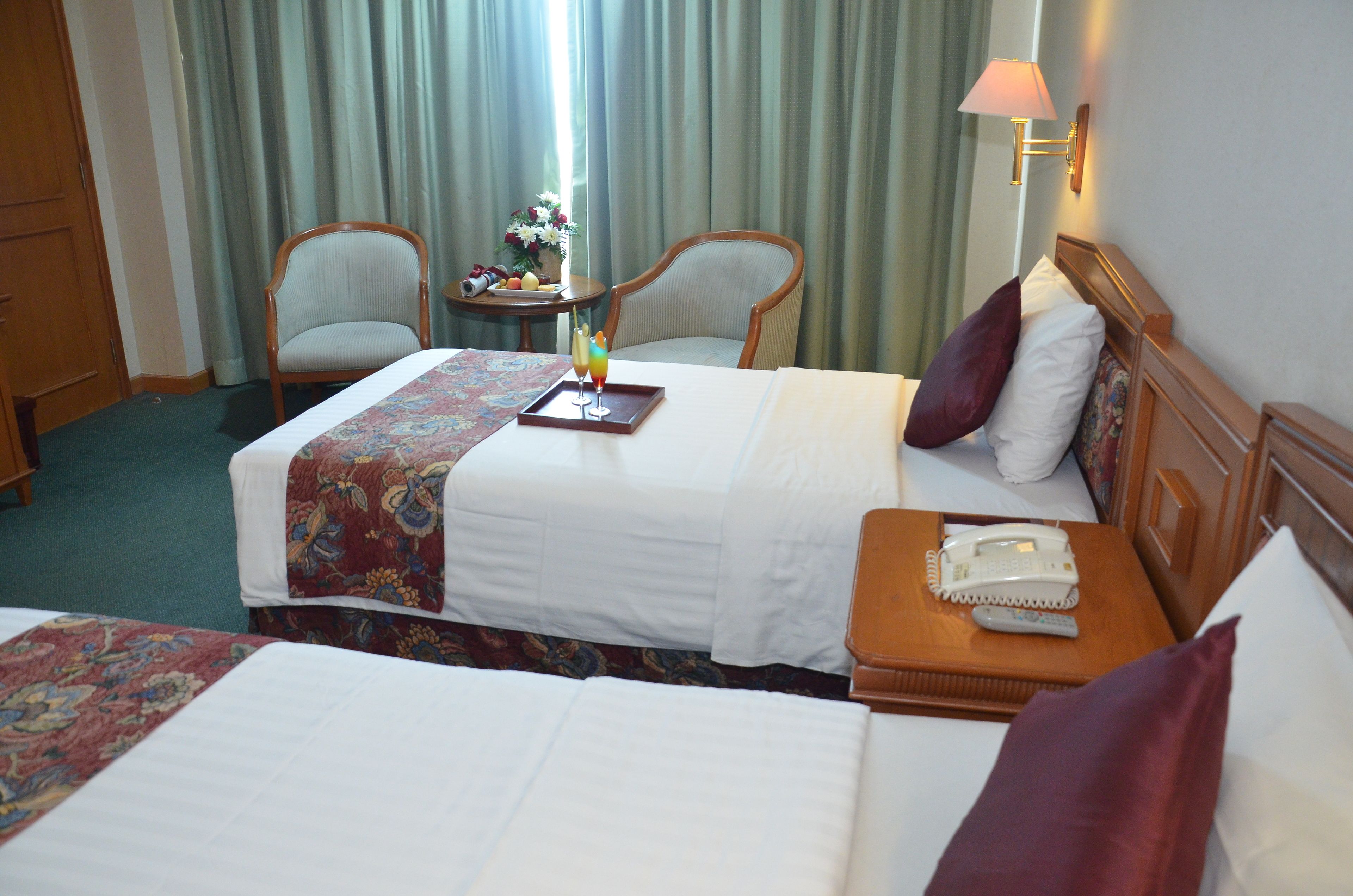 A hotel room with two beds, a bedside table with a lamp, a chair, and a small table with a vase of flowers.