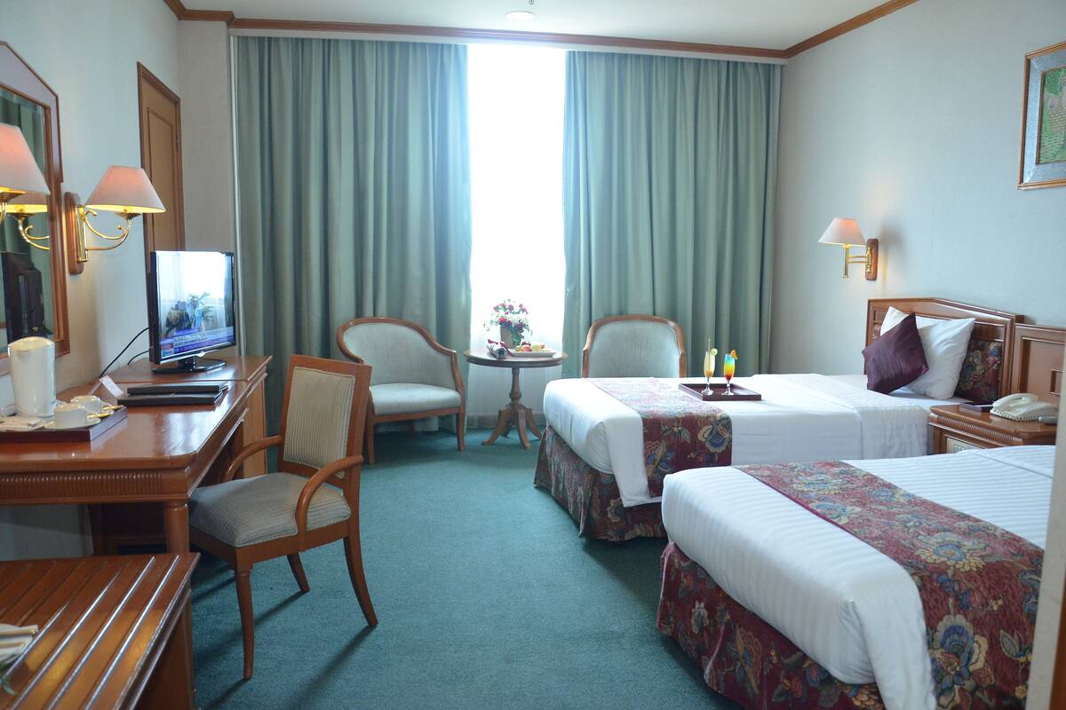 Une chambre d’hôtel avec deux lits, un bureau avec un ordinateur, une chaise, une petite table avec un vase et une fenêtre avec des rideaux.