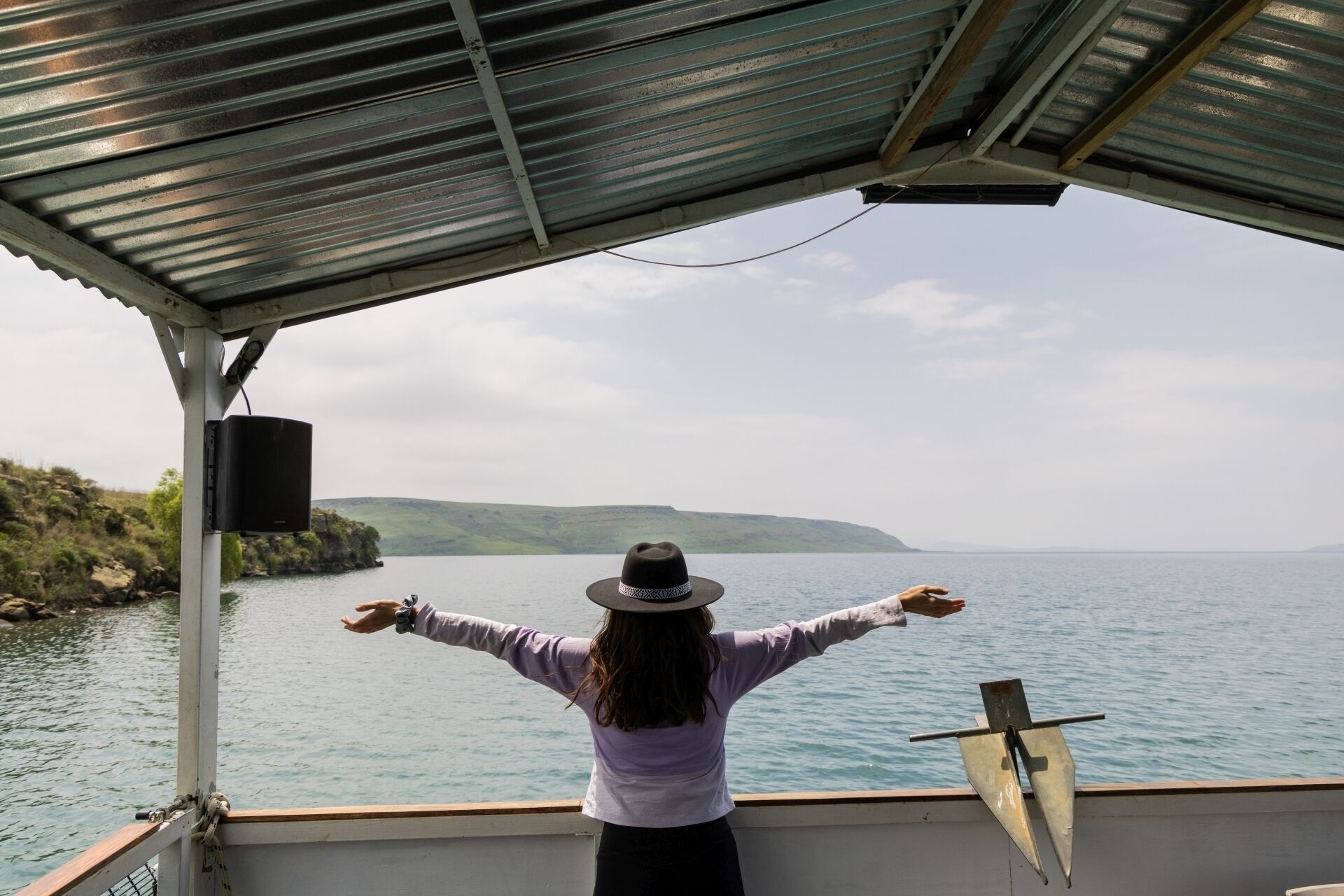 A person on a boat with a metal roof, looking out at a body of water and distant land.