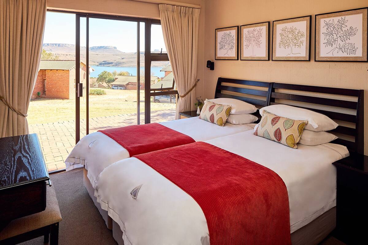 A double bed with a red and white bedspread, a wooden headboard, and a view of a landscape through a sliding door.