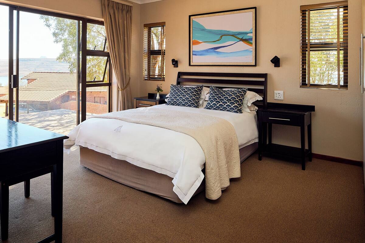 A bedroom with a large bed, a nightstand, a desk, and a window with a view of the outdoors.