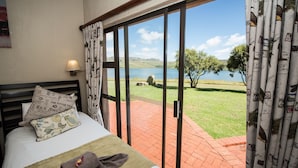 A bedroom with a bed, pillows, a towel, and a view of a lake and grassy area through large windows. - First Group Qwantani (Maluti a Phofung)