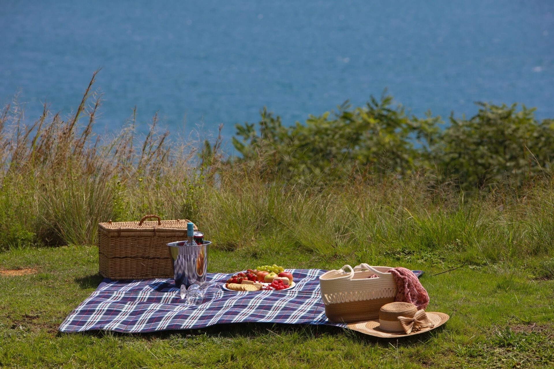 Un montaje para picnic con una manta, cesta de picnic y comida sobre césped cerca del agua.