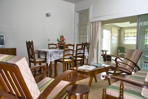 Un comedor con sillas de madera y una mesa, un televisor pequeño en un mueble, y una ventana con cortinas.