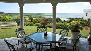 A covered patio with a glass table and chairs, overlooking a lush landscape and the sea. - Castles in Paradise (Vieux Fort)