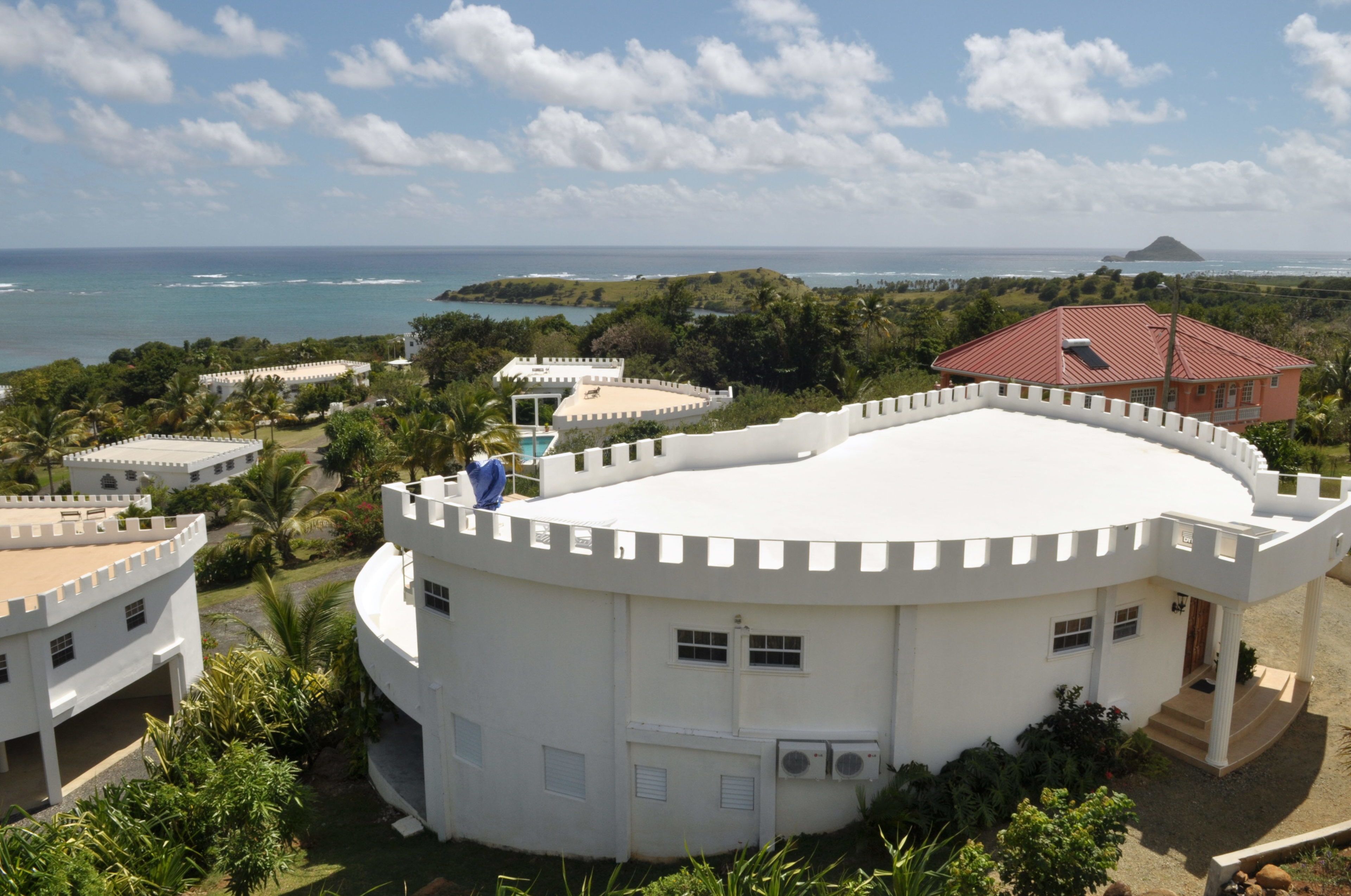 A white, circular building with a red-roofed structure nearby, surrounded by greenery and overlooking a coastal area.