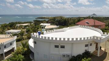 A white, circular building with a red-roofed structure nearby, surrounded by greenery and overlooking a coastal area.