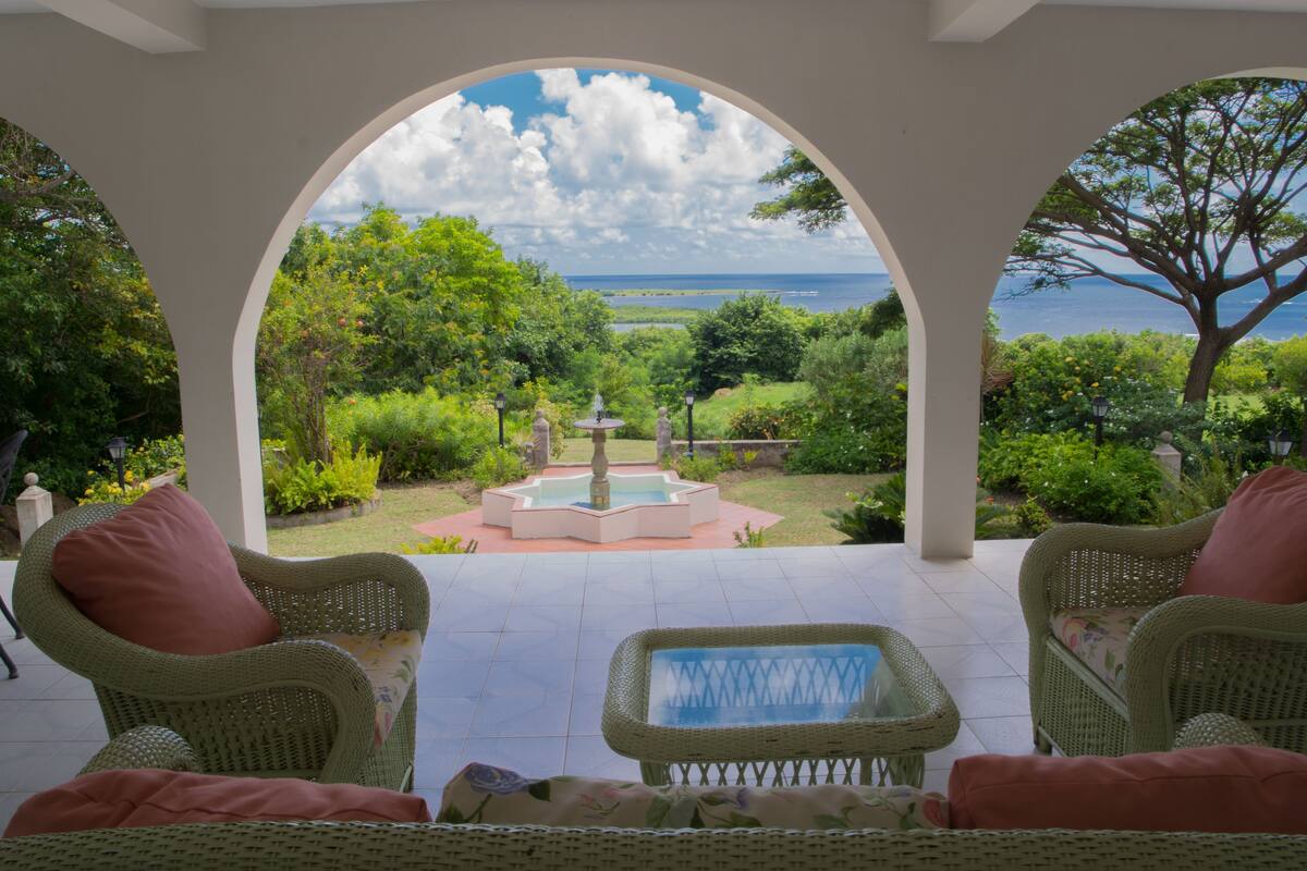 A covered patio with wicker furniture and a view of a garden and ocean.