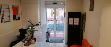Zona de recepción con mostrador, computadora y planta. Se ve una puerta que da a un balcón con alfombra roja.