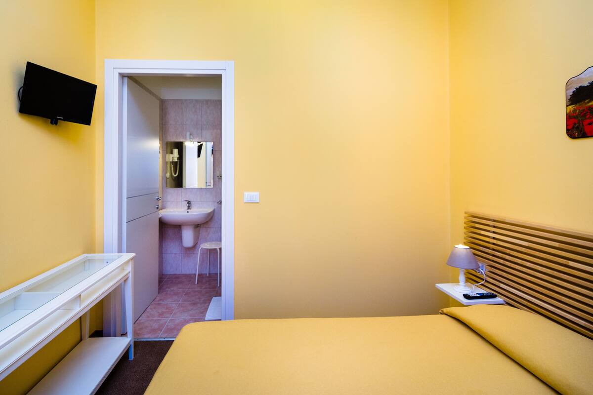 A compact hotel room with a bed, bedside table, lamp, TV, and a bathroom with a sink and mirror.