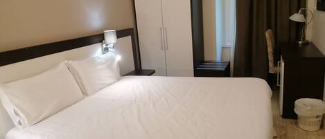 A hotel room with a large bed, a nightstand with a lamp, a wardrobe, and a view of a window with curtains.