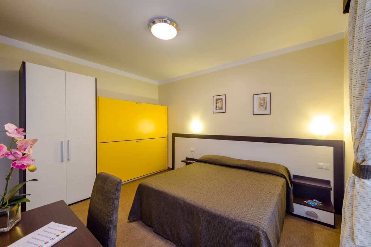A hotel room with a bed, a desk with a chair, a yellow wardrobe, and two framed pictures on the wall.