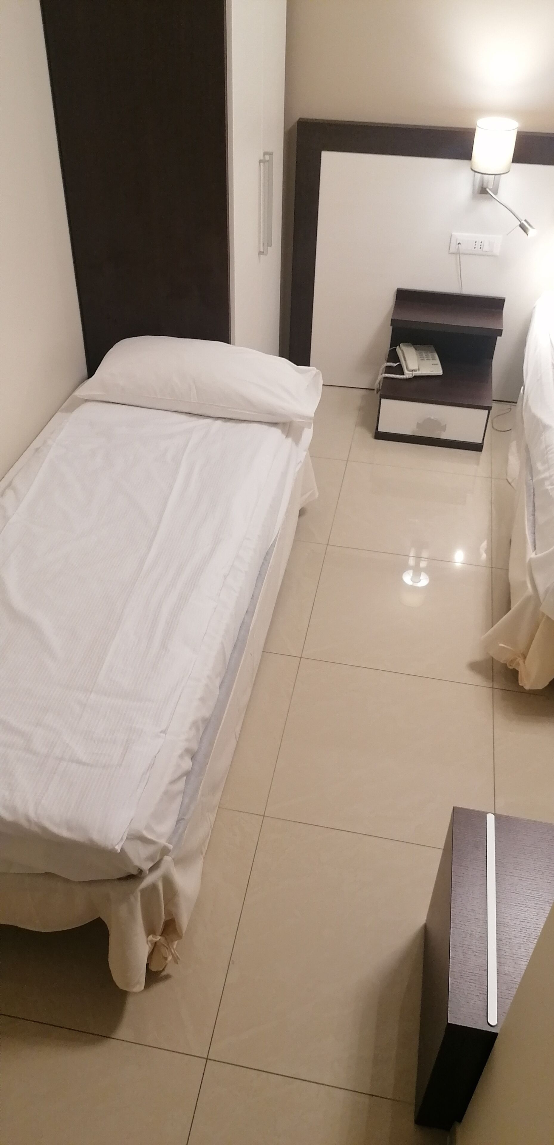 A hotel room with a bed, a bedside table with a phone, and a wall-mounted light.