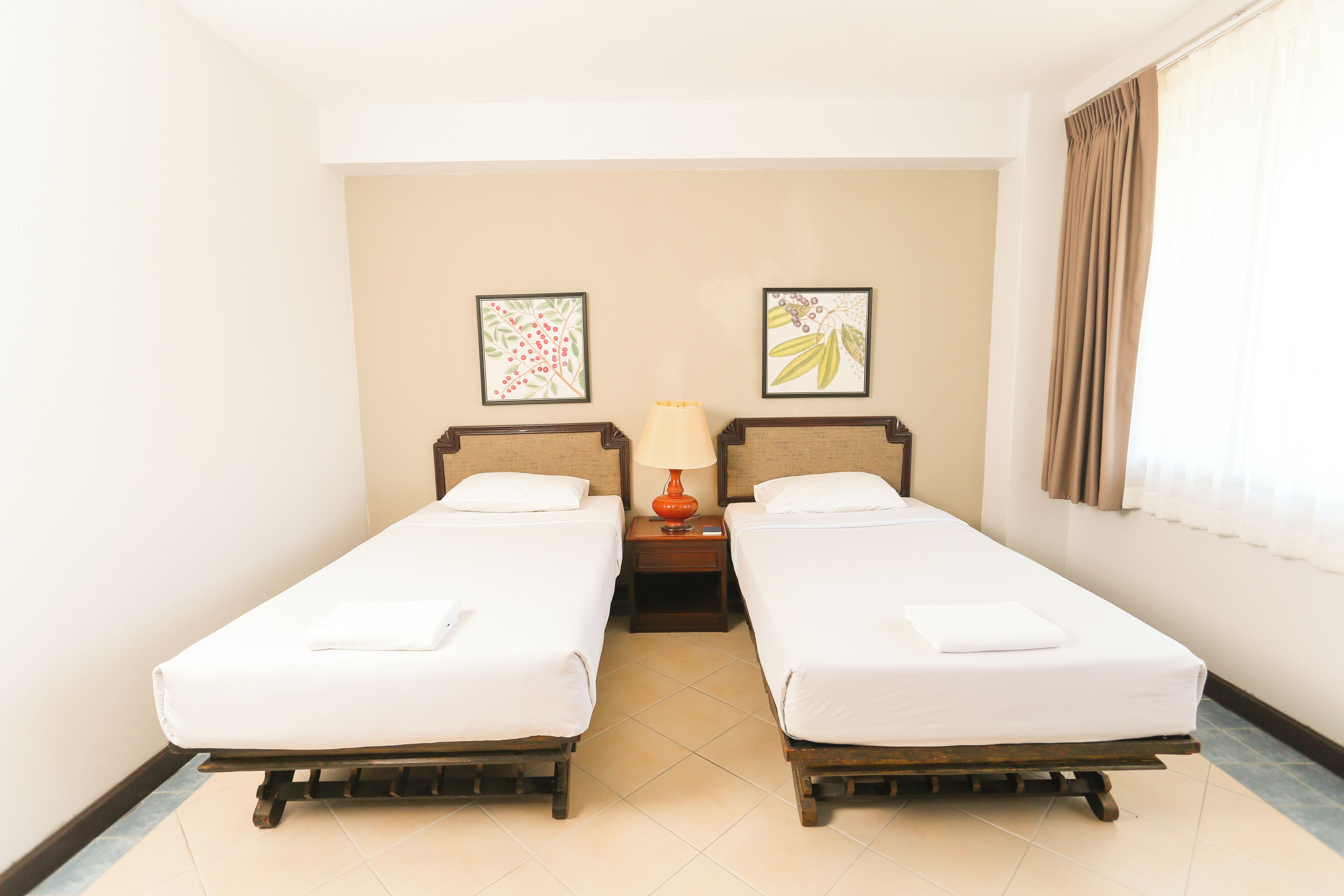 Habitación de hotel con dos camas individuales, una mesita de noche con lámpara y dos cuadros en la pared.