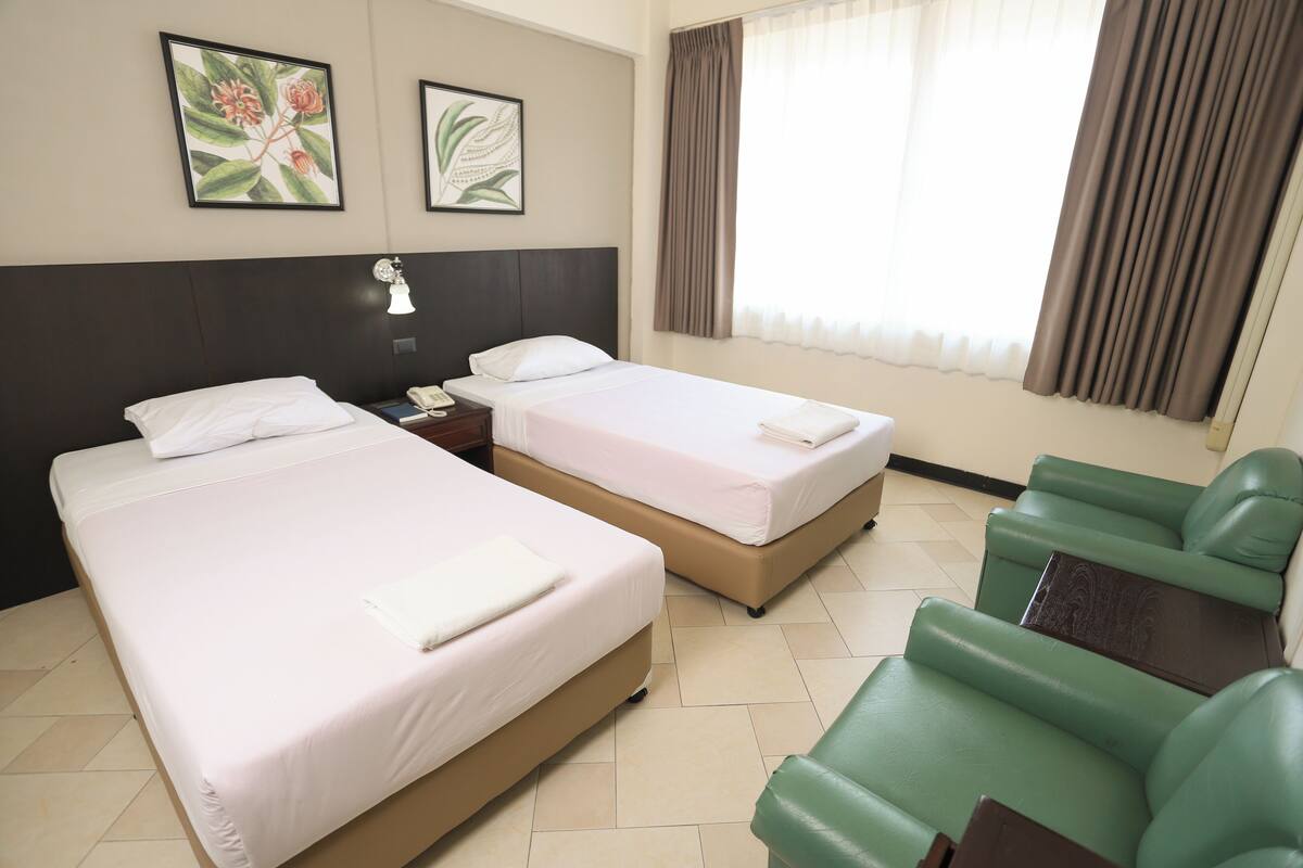A hotel room with two beds, a green sofa, a nightstand with a lamp, and two framed floral artworks on the wall.