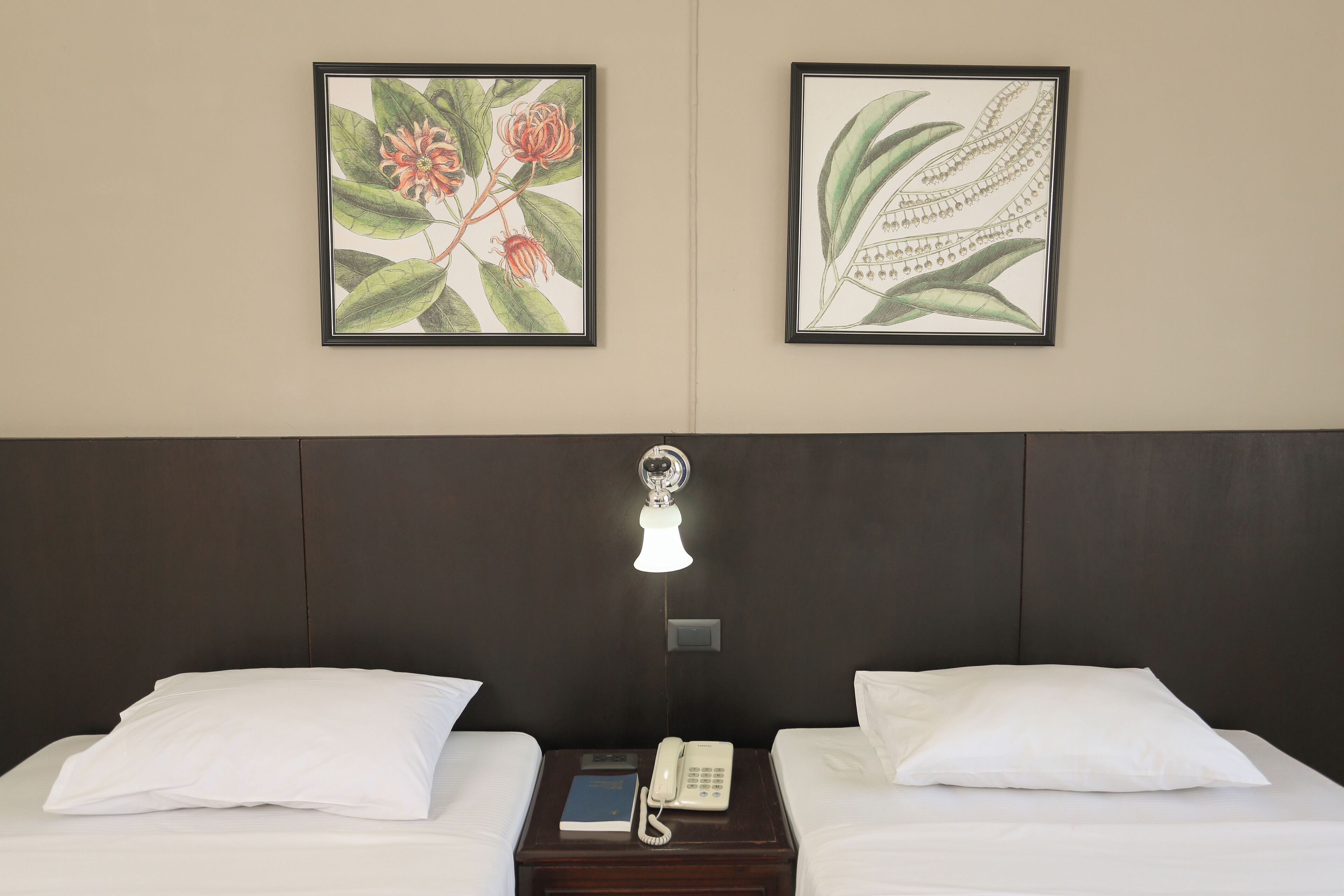 A hotel room with two beds, a nightstand with a telephone, and framed botanical artwork on the wall.