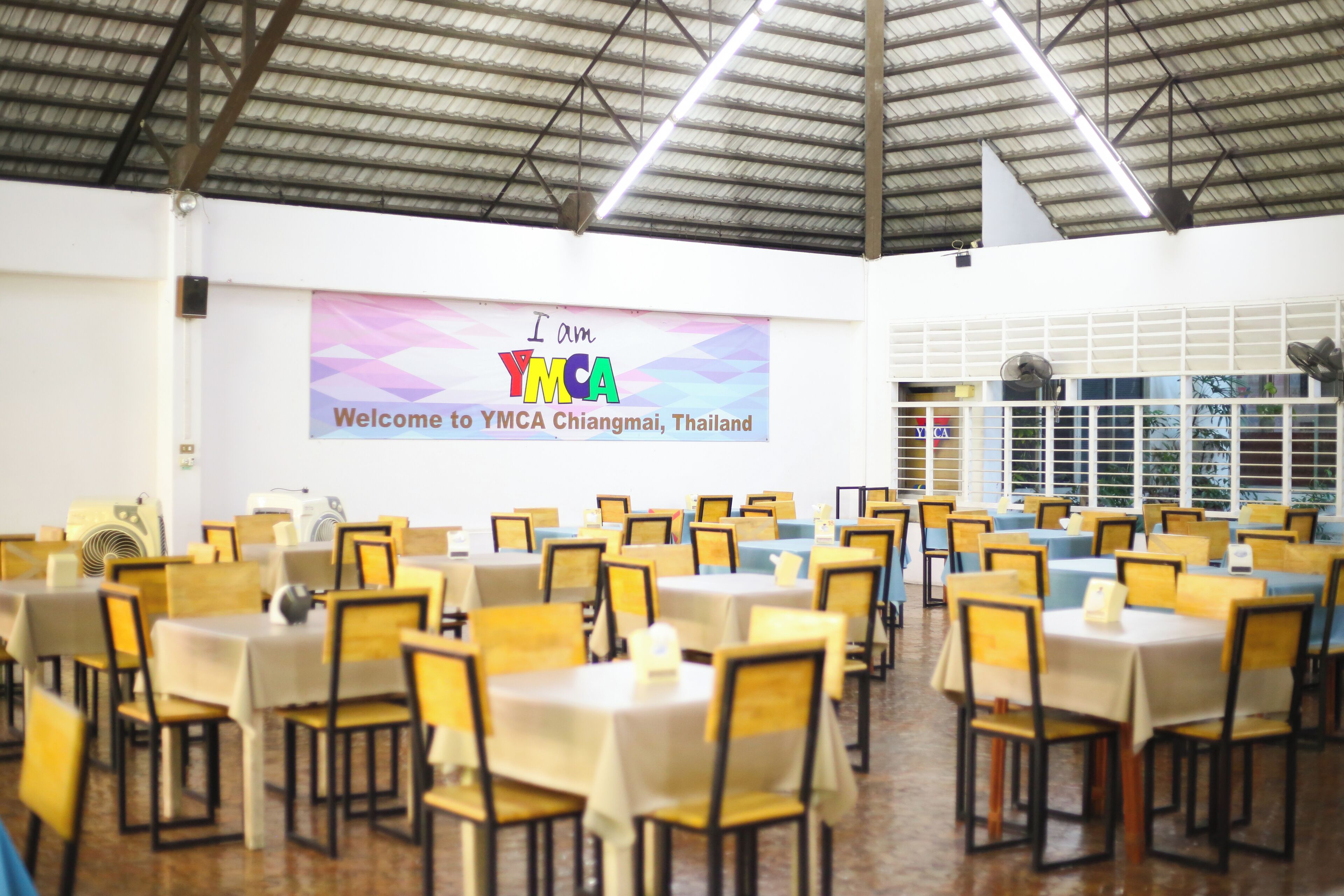 Een congrescentrum met tafels en stoelen, klaar voor een evenement. Een spandoek met 'I am YMCA Chiangmai, Thailand' hangt aan de muur.