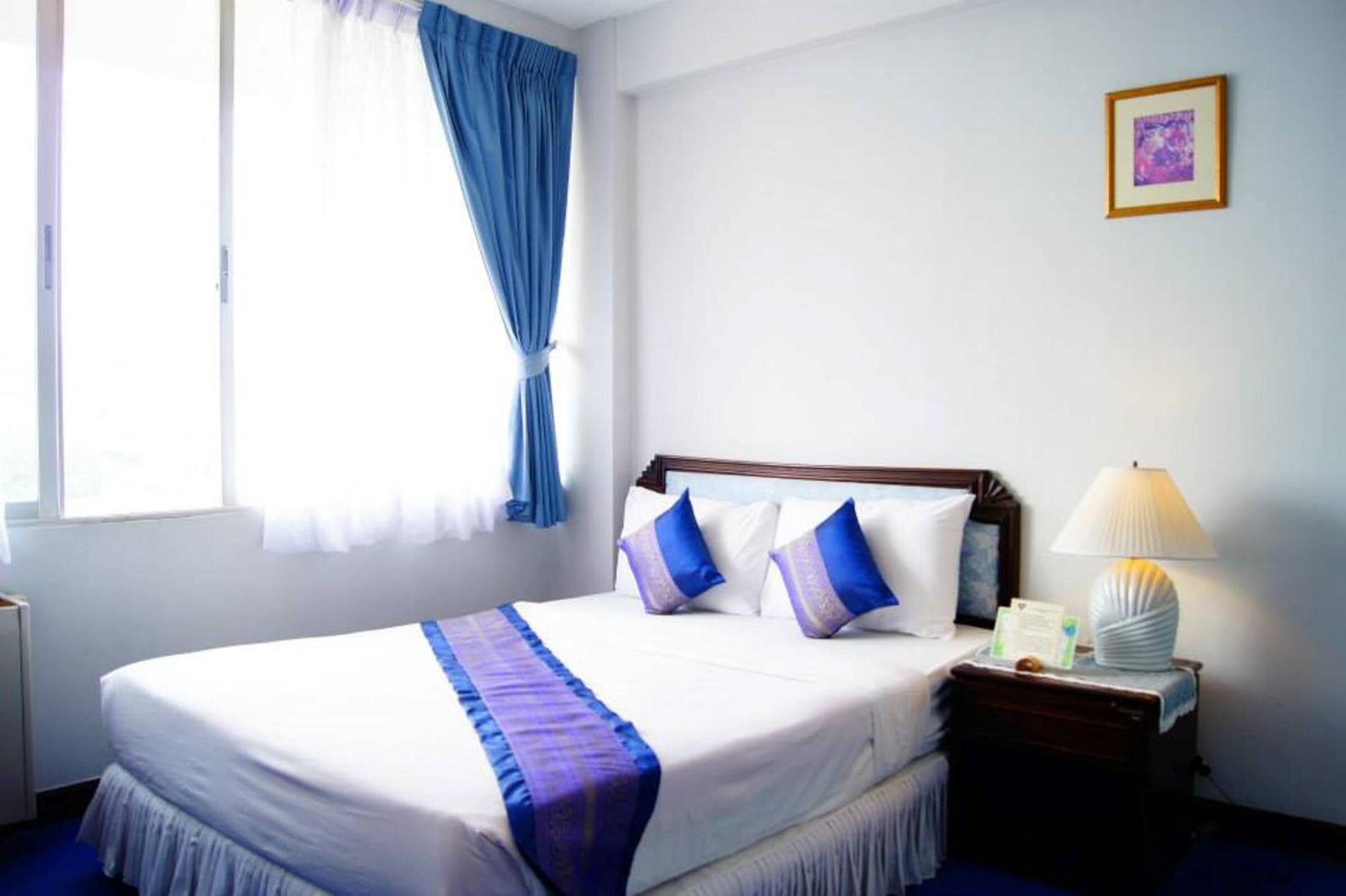 Habitación de hotel con una cama grande, una mesita de noche con lámpara, un cuadro enmarcado en la pared y una ventana con cortinas azules.