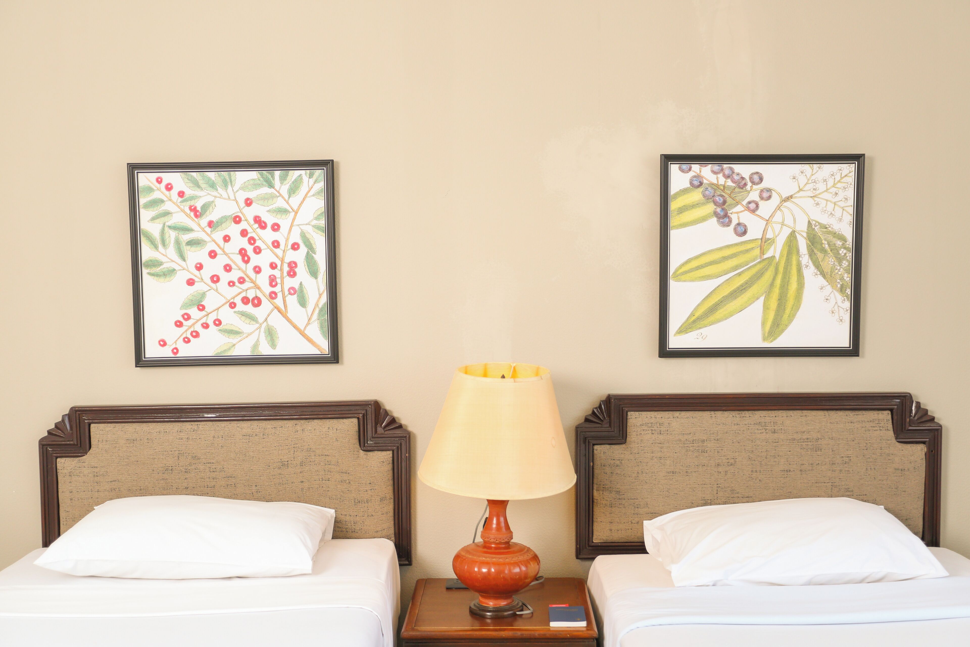 Habitación de hotel con dos camas, una mesita de noche con lámpara y dos impresiones enmarcadas con motivos botánicos en la pared.