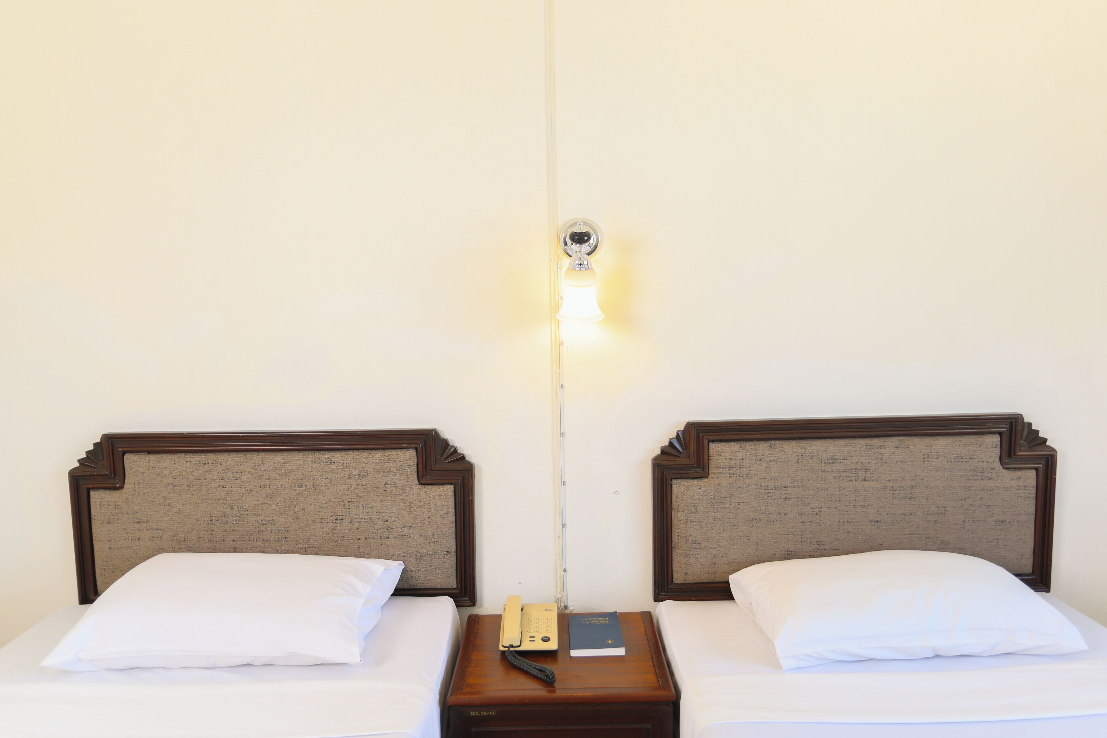 Une chambre d’hôtel avec deux lits simples, chacun doté d’une tête de lit en bois et de draps blancs. On y trouve une table de chevet avec un téléphone et un livre.