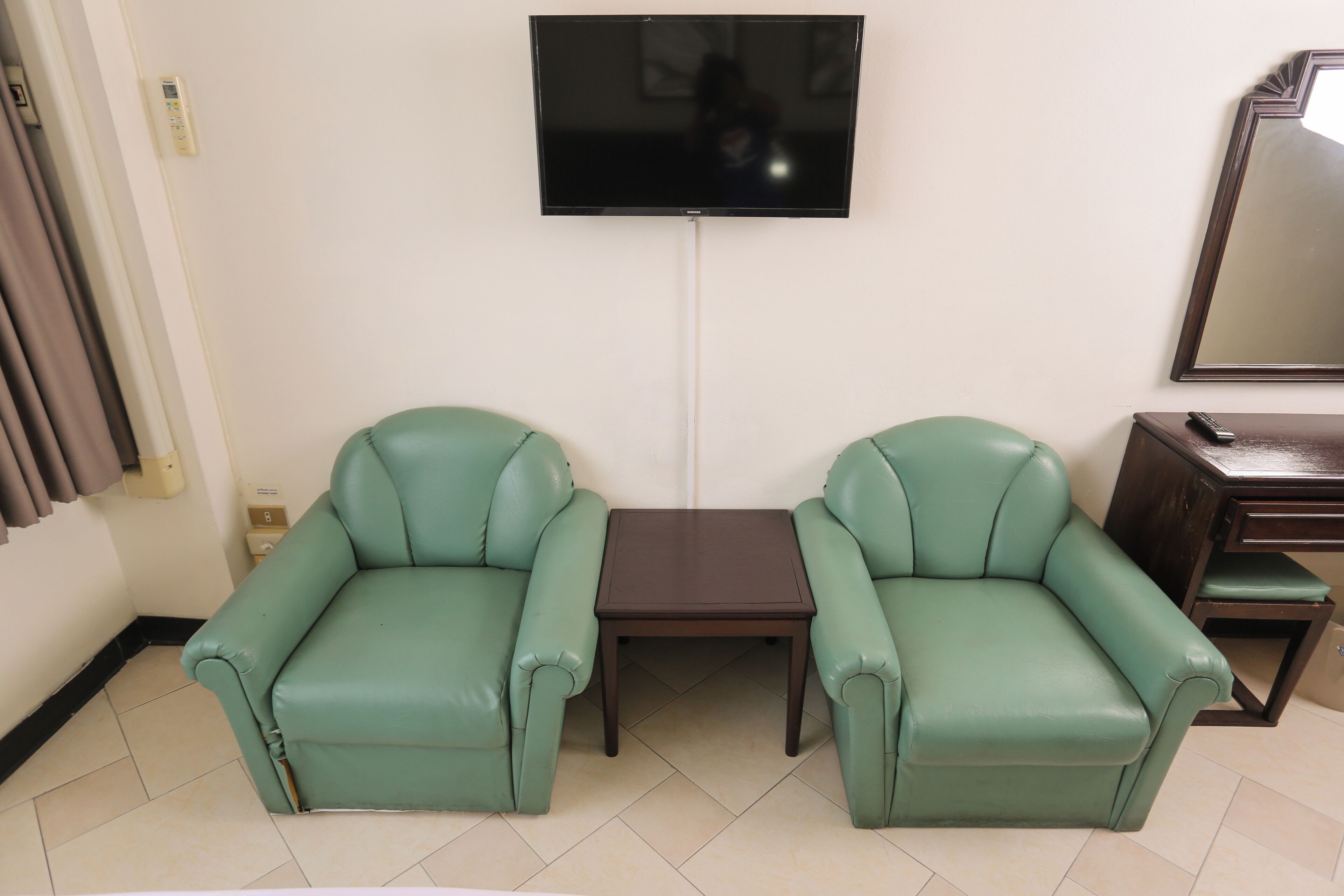 A room with two green leather armchairs, a small table, a TV mounted on the wall, a mirror, and a wooden cabinet.