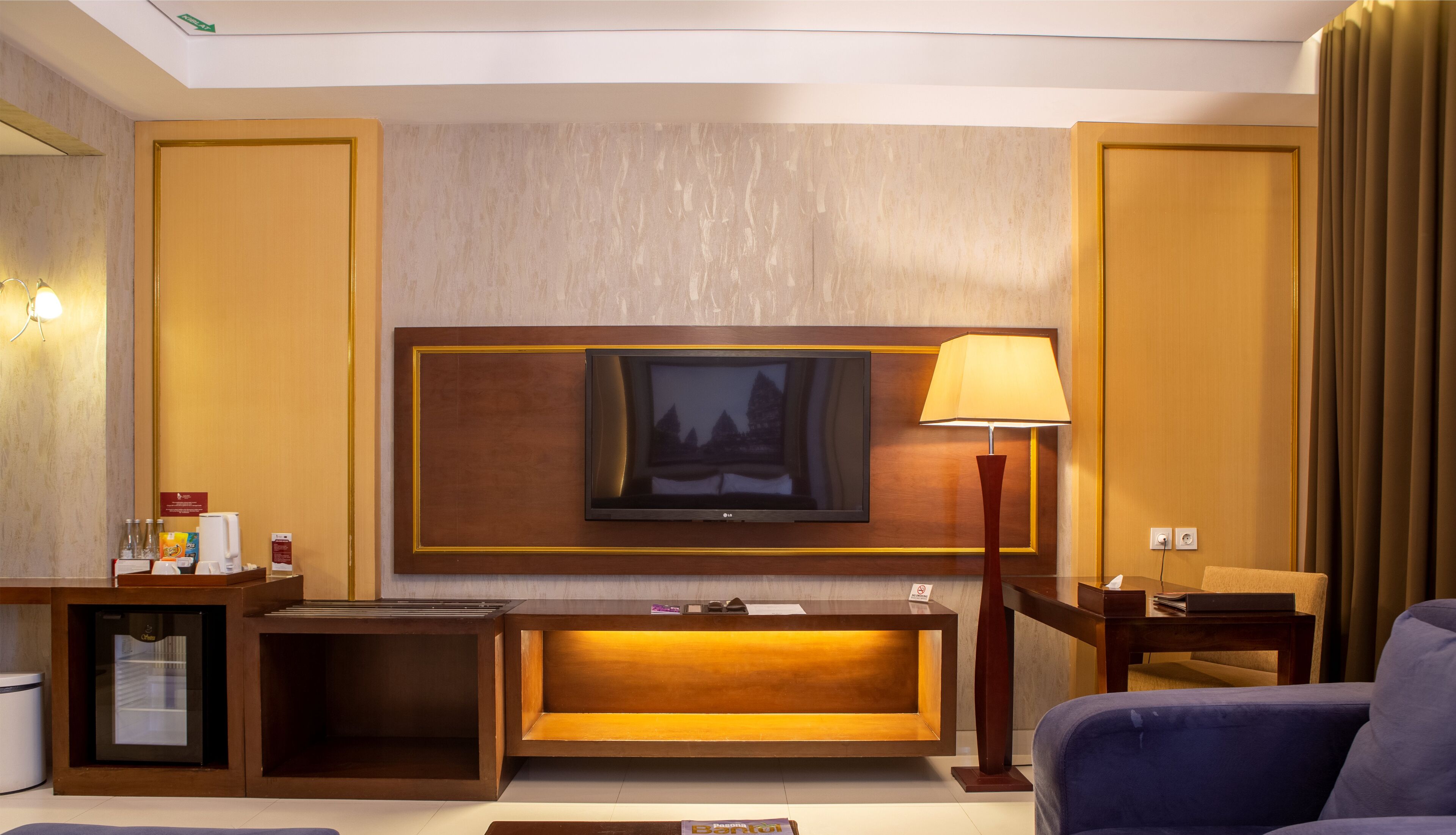 A hotel room with a large flat-screen TV mounted on the wall, a wooden console table, a blue sofa, and a lamp.
