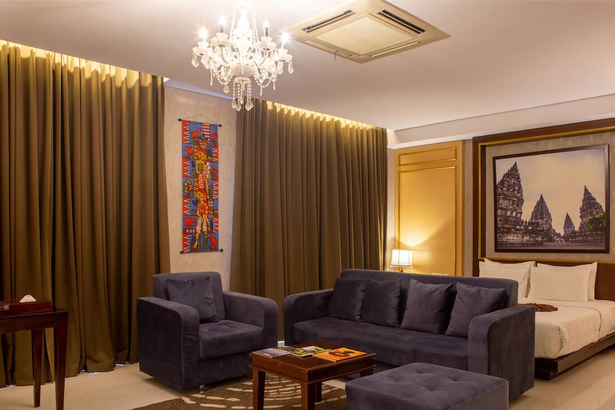 Hotelkamer met een groot bed, twee fauteuils, een bijzettafeltje, een kroonluchter en een schilderij van een tempel.