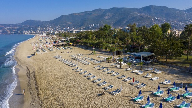 A beach with rows of sun loungers and umbrellas, a lifeguard tower, and a backdrop of mountains and a city. - Aslan City Hotel (Alanya)
